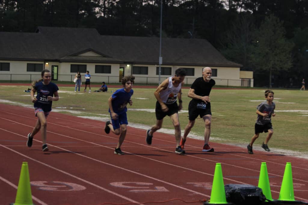 Four runners of various ages sprint along a red track at a Florida track club field.