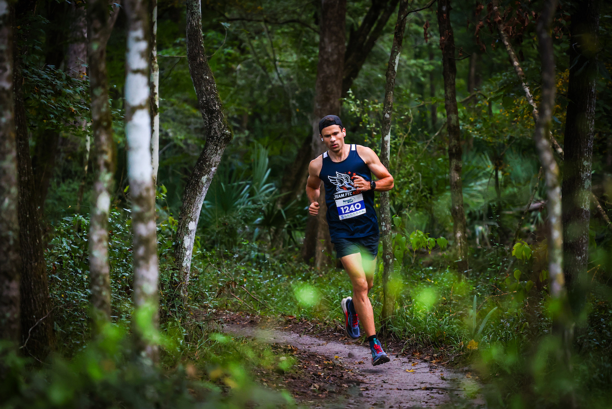 A Florida Track Club runner wearing a tank top and race bib 1240 runs on a wooded trail.
