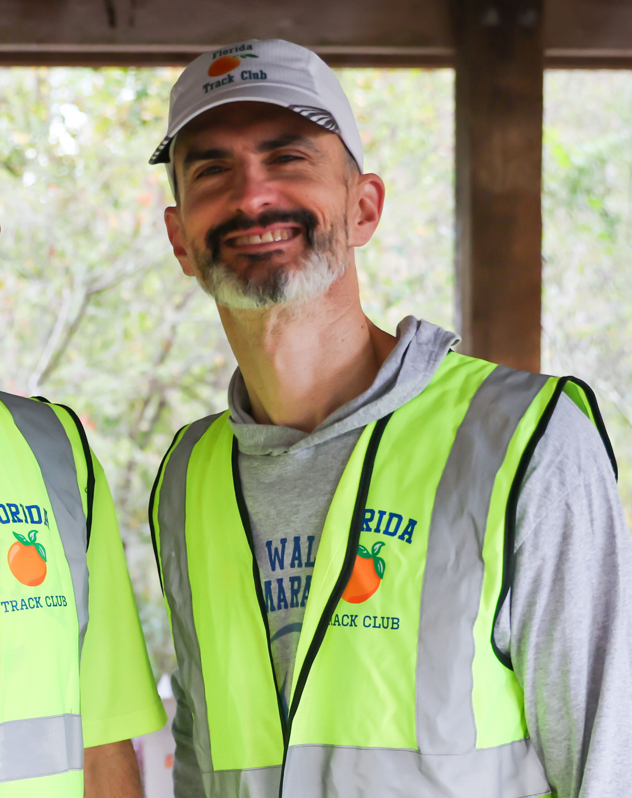 A smiling man wearing a Florida Track Club baseball cap and a neon yellow safety vest over a grey sweatshirt, representing the Gainesville running club.