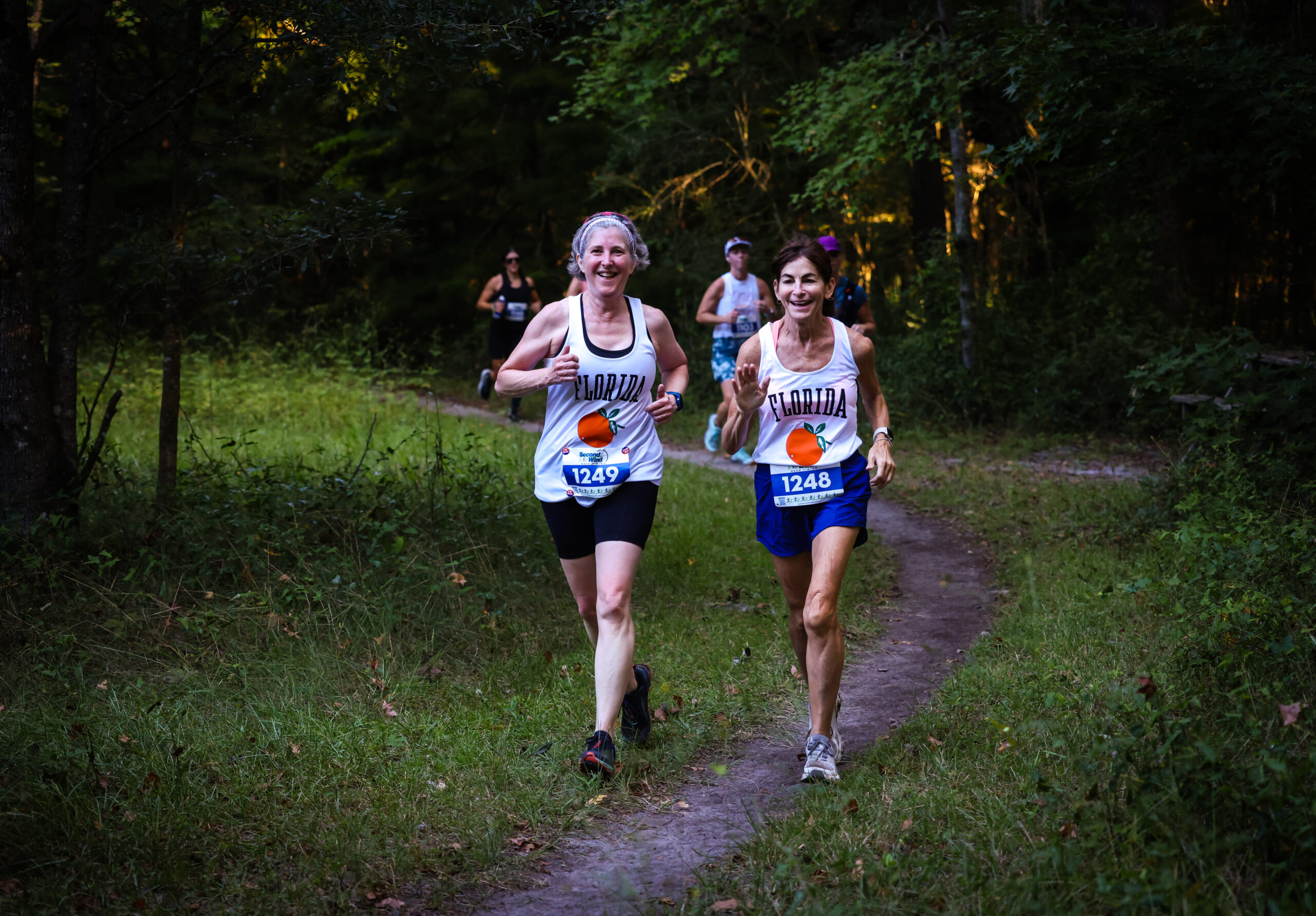 Two runners from the Florida Track Club wearing race bibs 1249 and 1248 jog along a wooded trail during a race.