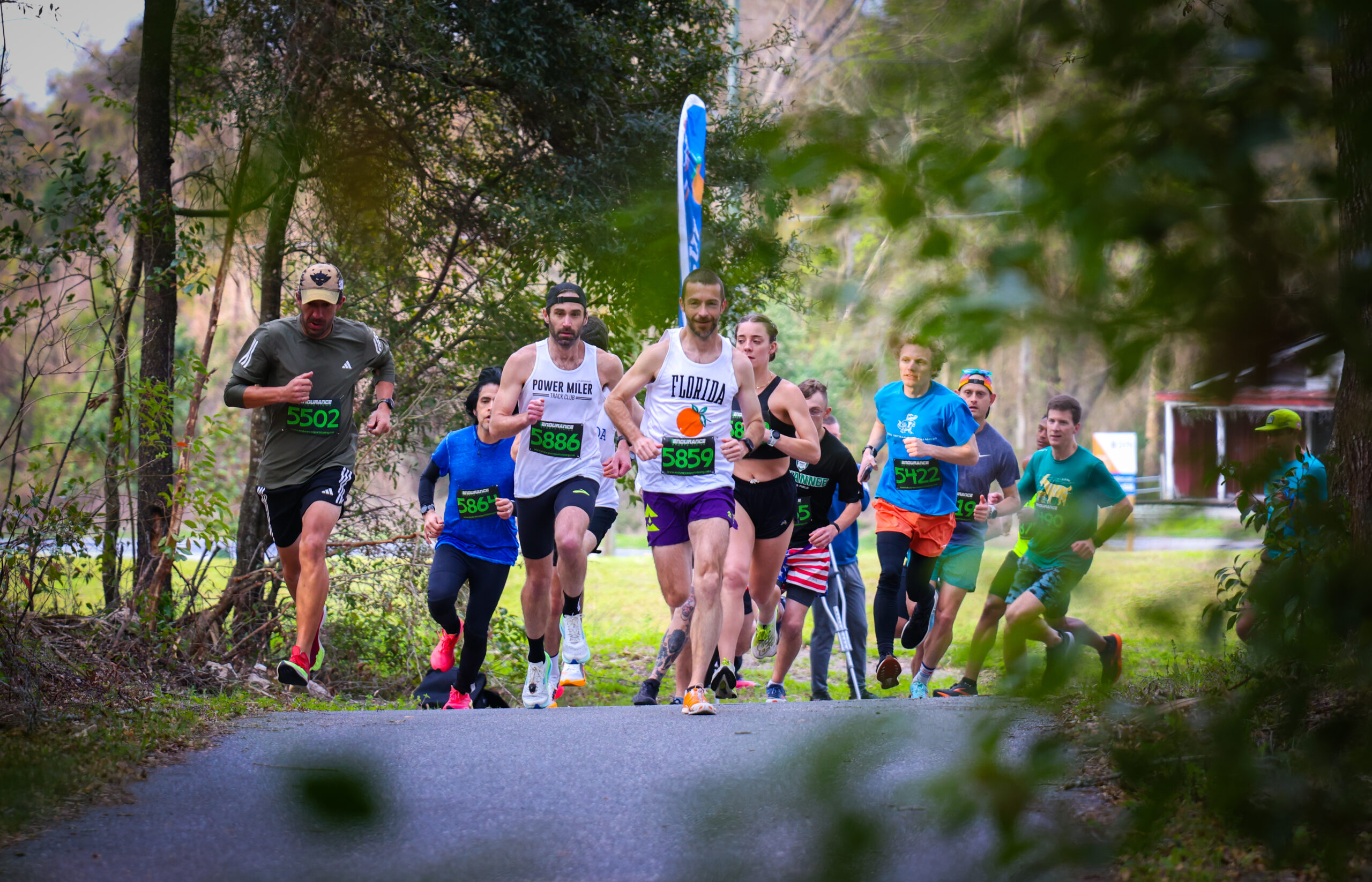 A group from the Gainesville Running Club participates in a road race on a tree-lined path.