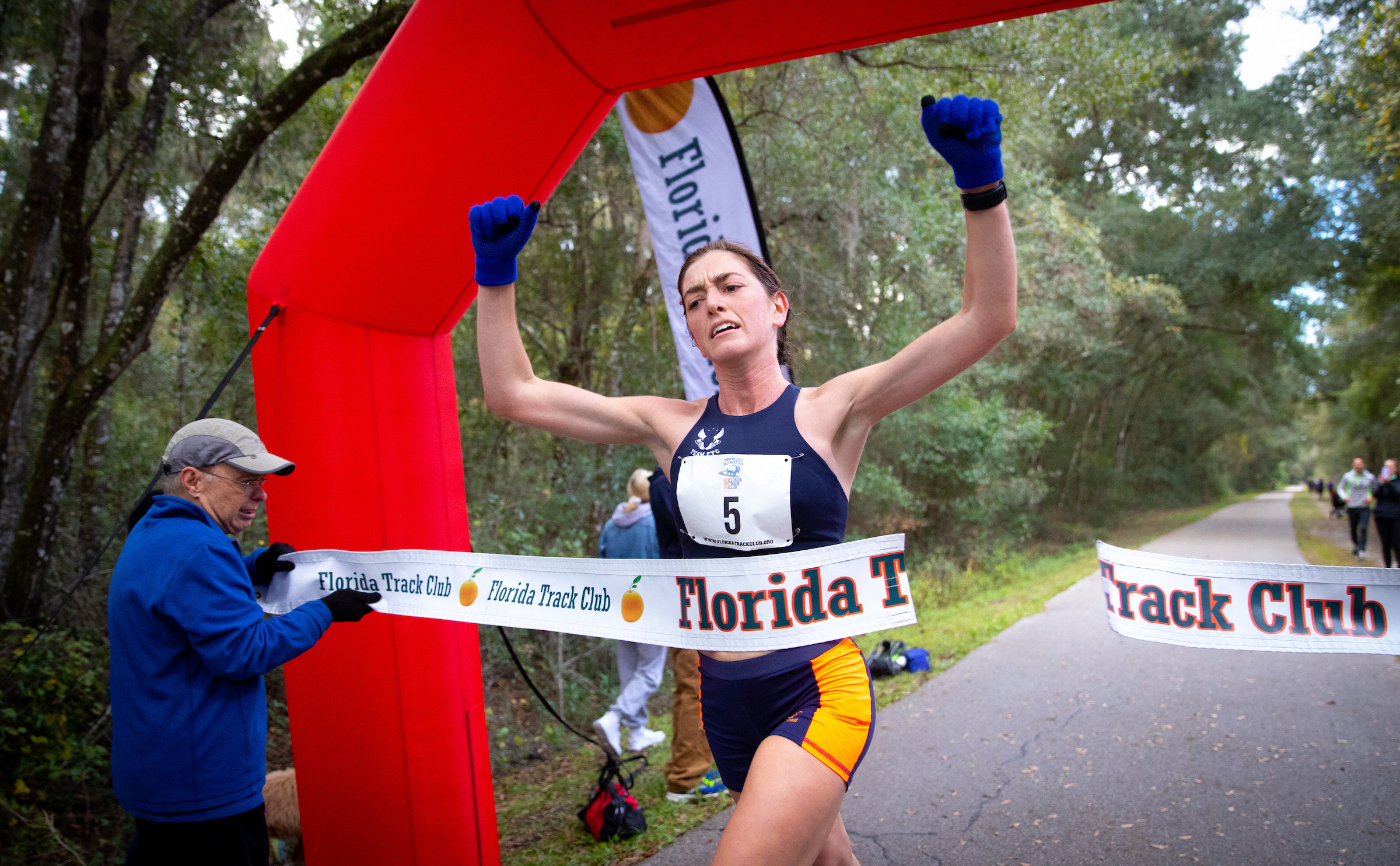 A female Florida Track Club runner wearing a race bib with the number 5 crosses a finish line held by an official.