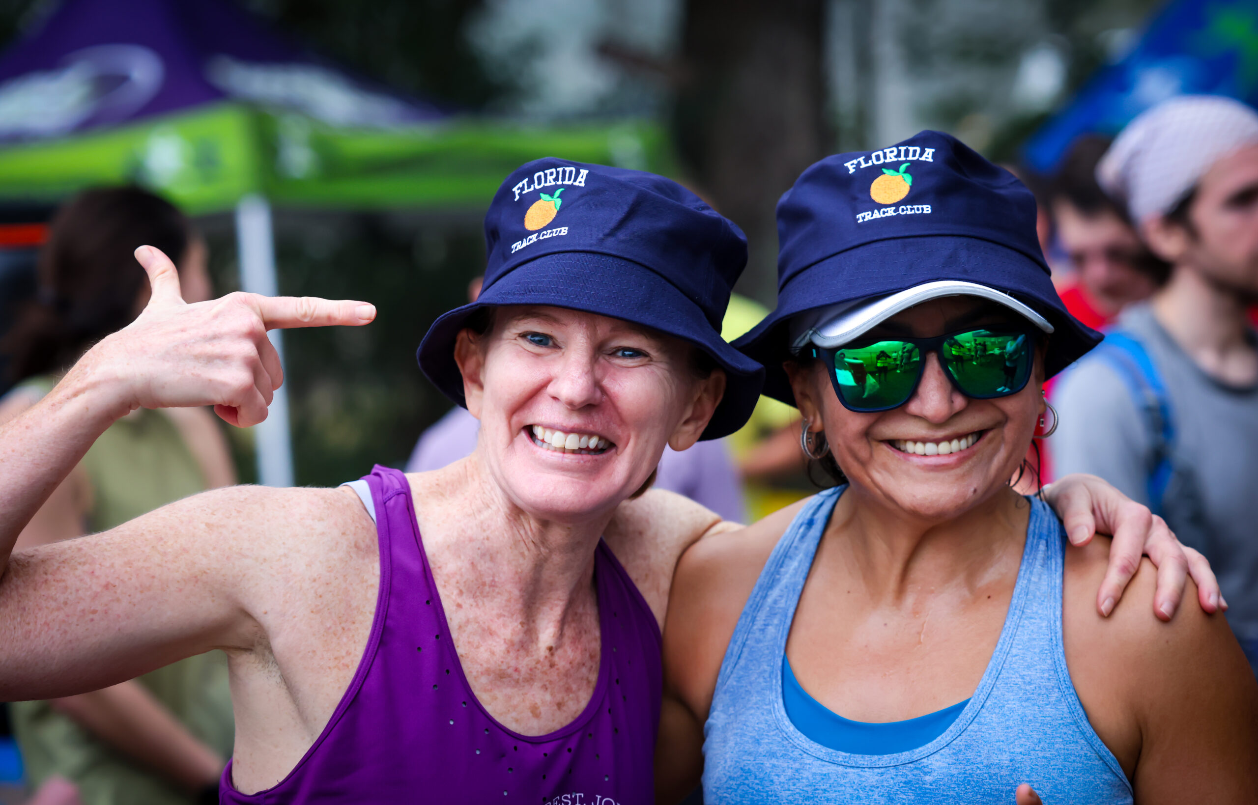 Two women wearing blue Florida Track Club bucket hats from the Gainesville running club smiling at the camera.