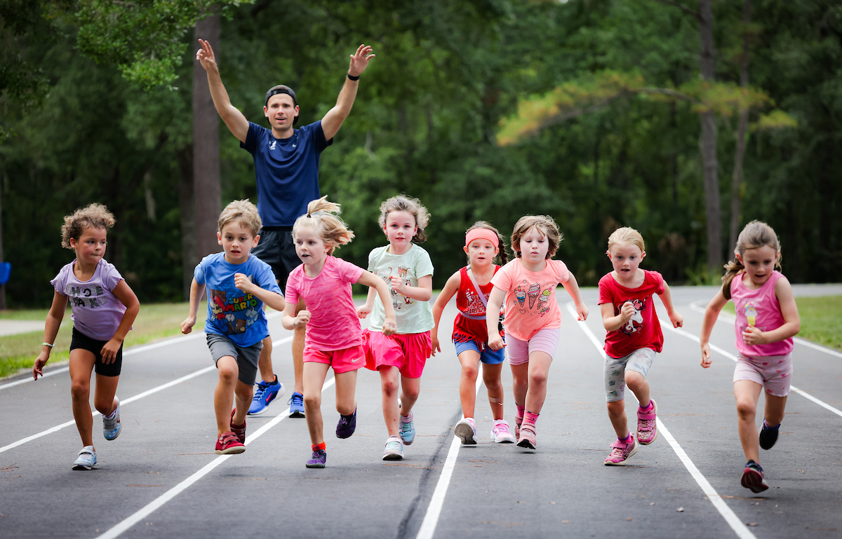 A man cheers behind a group of seven young children from a Gainesville running club as they run across a track.