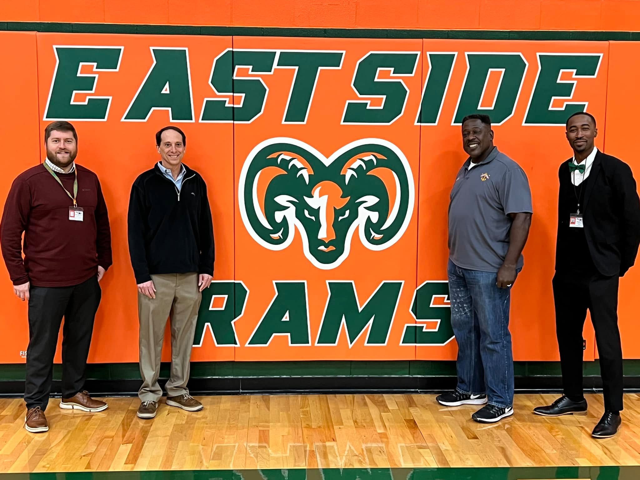 Four men stand in a gymnasium in front of a wall painted with the Eastside Rams logo and team name, representing the Florida Track Club.