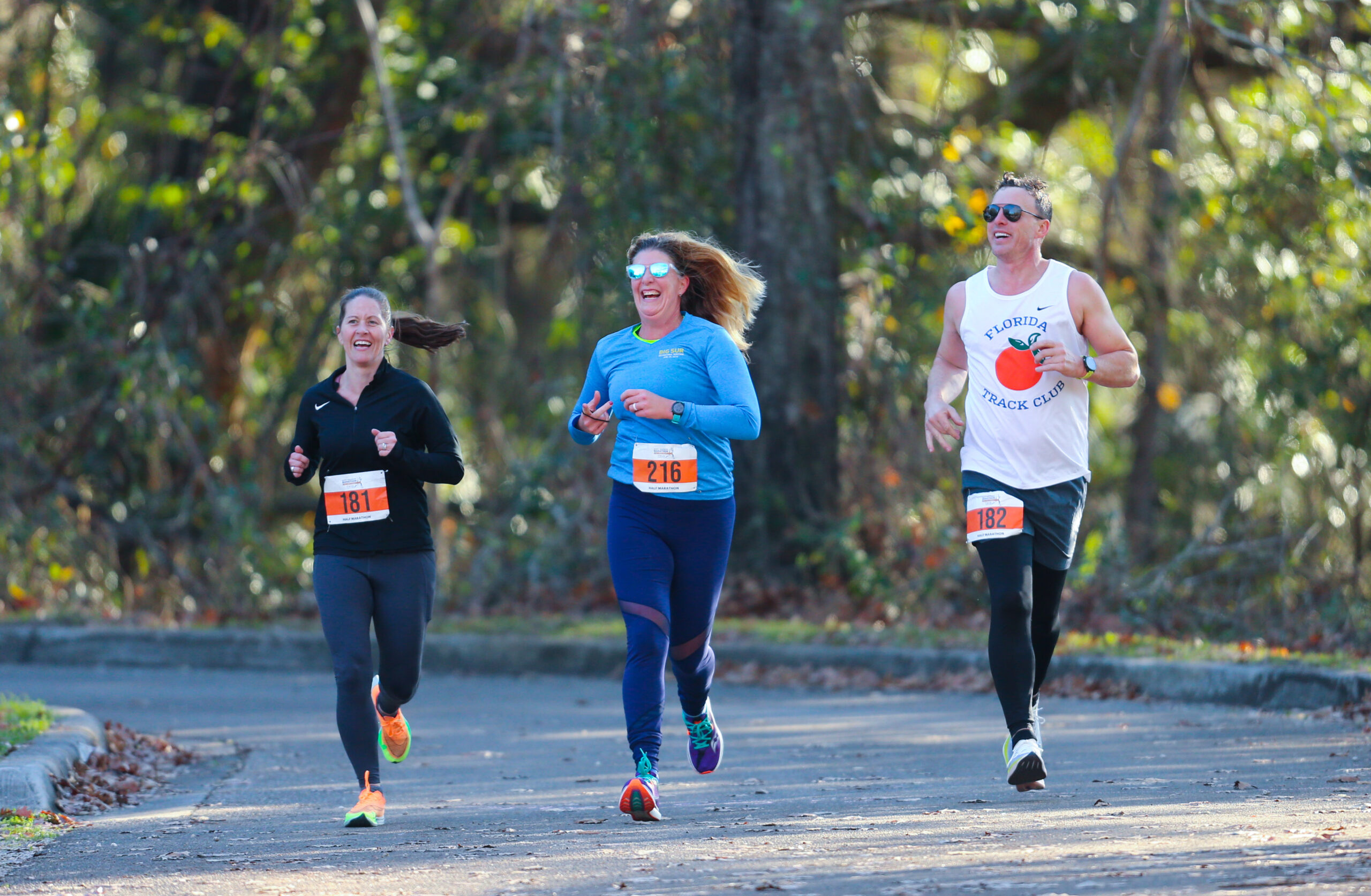 Three runners from the Florida Track Club laughing while jogging along a paved path.