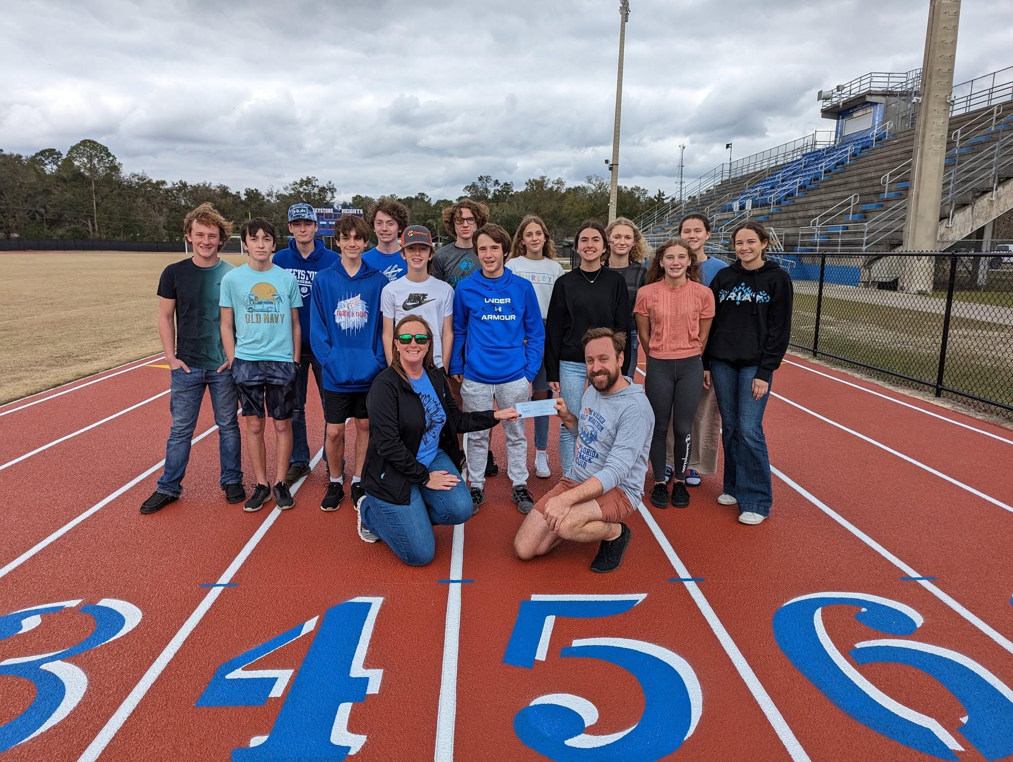 A group of teenagers and two adults stand on a running track, with one adult and one student holding a ceremonial check for the Gainesville Running Club.