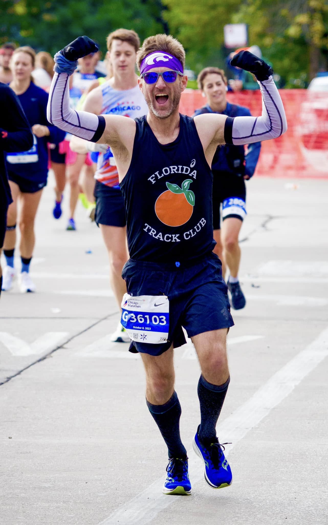 A runner wearing a Florida Track Club tank top and a purple headband celebrates with arms raised while crossing the finish line at the Chicago Marathon.