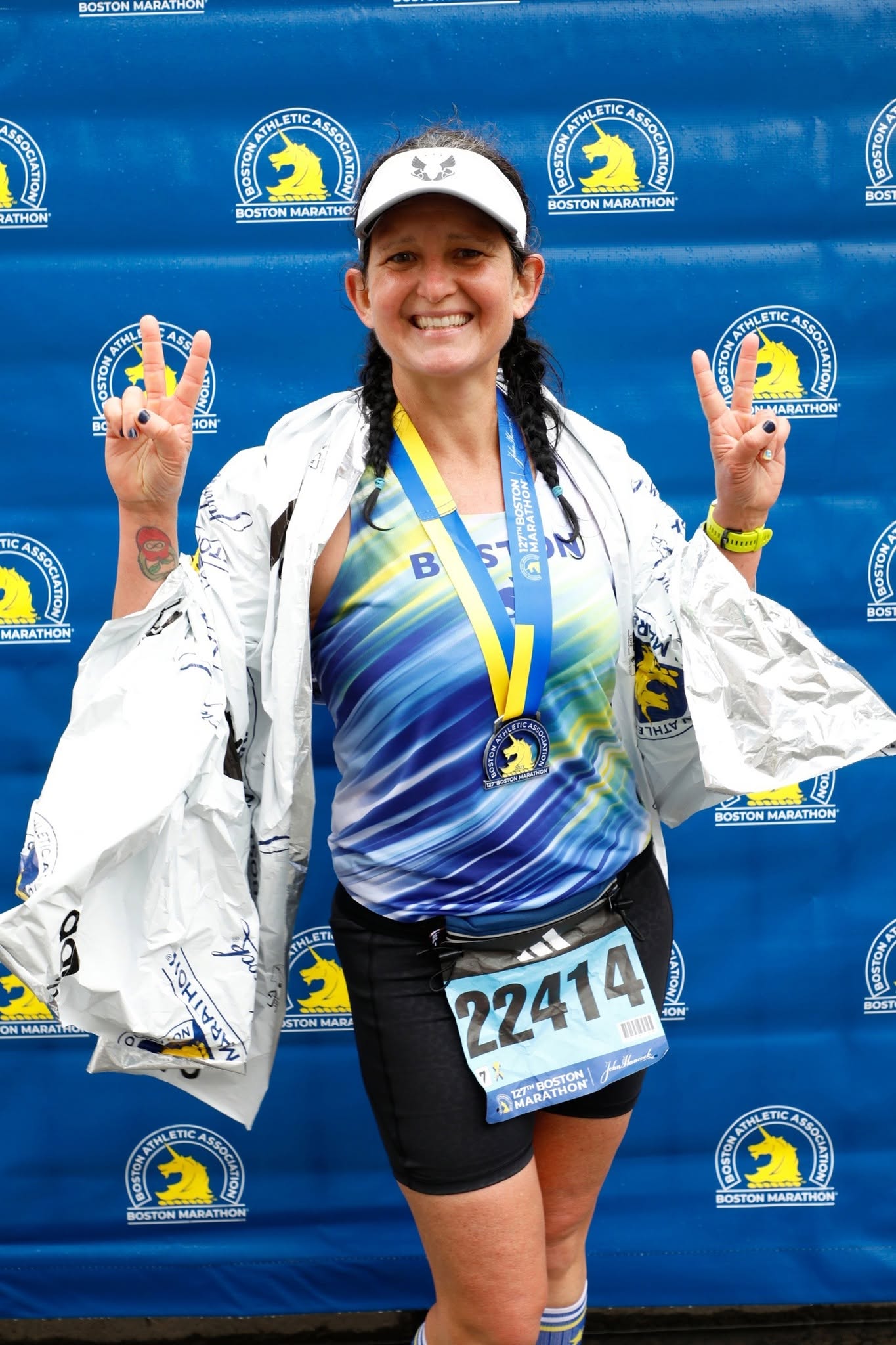 A smiling Gainesville Running Club member wearing a Boston Marathon medal and foil blanket makes peace signs with both hands in front of a blue Boston Marathon backdrop.