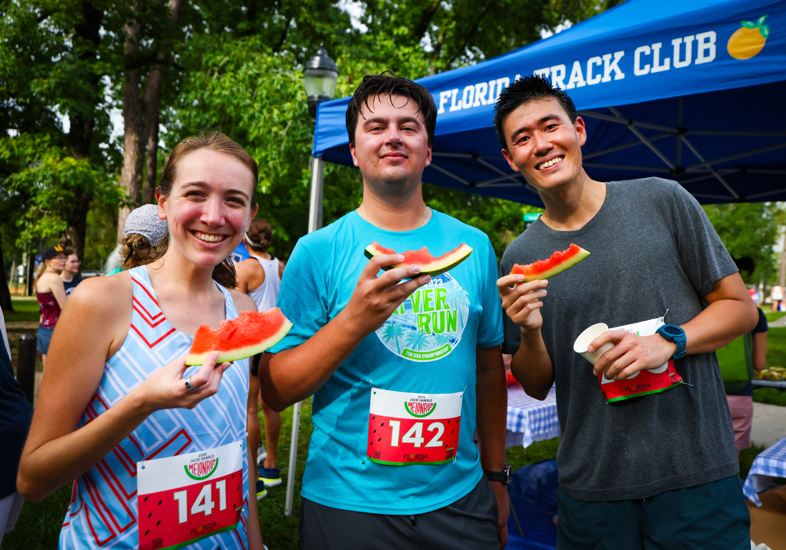 Three runners in race bibs stand in front of a Florida Track Club tent, each holding a slice of watermelon.