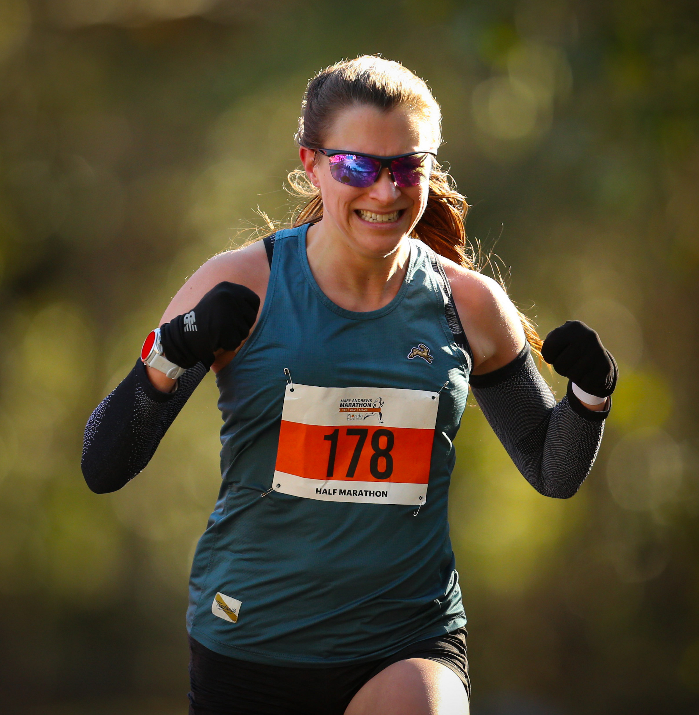 A determined Gainesville Running Club member races in a half marathon, wearing a teal tank top with bib number 178, sunglasses, and arm sleeves.