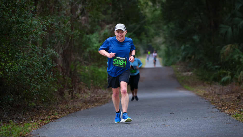 A man wearing a blue shirt and race bib number 5884 running on a paved path during a Gainesville Running Club event.