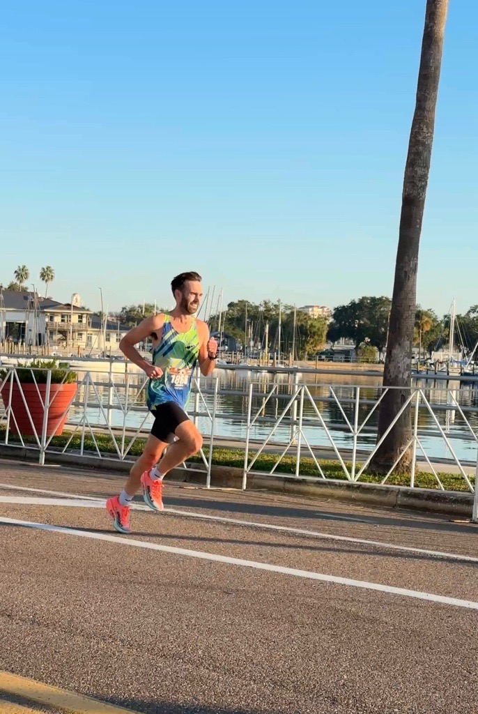 A male runner in a colorful tank top and black shorts running along a waterfront path with the Gainesville Running Club.