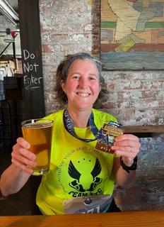 A smiling woman in a neon yellow running shirt from the Gainesville Run Club holds a glass of beer and a half-marathon finisher medal.
